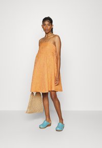 Vestido de verão laranja com alças finas, tecido texturizado e forma expandida. Combinado com uma bolsa de palha e tamancos de madeira azuis.