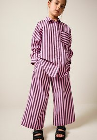Oversize-Hemd mit vertikalen pinken und schwarzen Streifen und passenden weit geschnittenen Hosen, mit Knopfleiste und einer Brusttasche. Schwarze Sandalen.