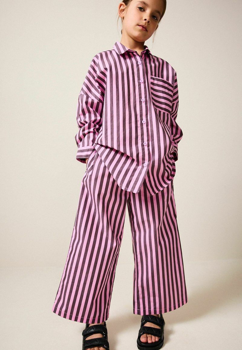 Oversize-Hemd mit vertikalen pinken und schwarzen Streifen und passenden weit geschnittenen Hosen, mit Knopfleiste und einer Brusttasche. Schwarze Sandalen.