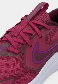 Nike sportiniai batai su tamsiai rožinės spalvos tinkleliu, tekstūruotomis detalėmis, violetiniais akcentais ir plokščiomis raišteliais. Balta padu su griovelių raštu.