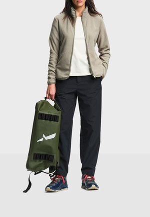 Kobieta w beżowej kurtce polarowej, czarnych spodniach i niebieskich trampkach trzymająca oliwkowozieloną torbę z białym logo na jasnym tle.