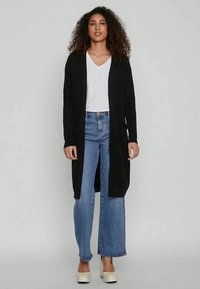 Svart lang cardigan laget av mykt stoff, åpen frontdesign, kombinert med en hvit V-hals skjorte og lyseblå vidbukser. Uformell stil.