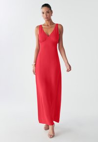 Pikk punane kleit, millel on lõhikuga V-kaelus, varrukateta disain ja sile kangas. Omaduseks on istuv ülakeha ja voolav seelik. Muster või kaunistused puuduvad.