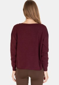 Donna con lunghi capelli castano chiaro che indossa un maglione largo di colore rosso scuro e pantaloni marroni, vista di spalle su sfondo bianco.