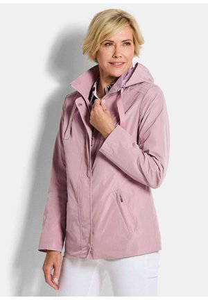 Blonde Frau, die die Kapuze einer hellrosa Zip-up-Jacke anpasst, trägt weiße Hose, steht vor einem weißen Hintergrund.