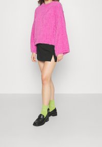 Luddig rosa tröja, svart minikjol med sidoflärp, och gröna knästrumpor som matchas med svarta loafers. Enkel bakgrund.