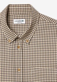 Koszula w kratę w odcieniach beżu i granatu; posiada klasyczny kołnierzyk, regularny krój oraz drewniane guziki. Widoczne logo Lacoste.
