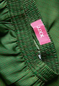 Grön elastisk midja med samlad textur och horisontella ränder i varierande nyanser av grönt. Rosa etikett med texten "JXX".