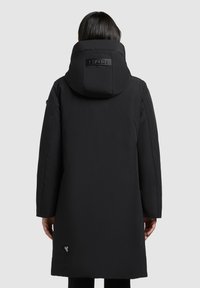 Isik kannab pikka musta kapuutsiga mantlit, millel on polstriga seljaosa ja brändipatch allääres, seistes ühetaolise tausta ees.