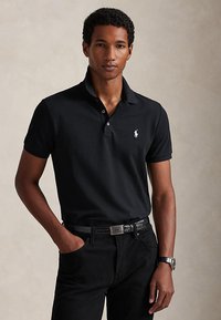 Homme portant un polo noir à manches courtes, un pantalon noir avec ceinture et une montre-bracelet, debout avec une main dans la poche sur un fond uni.