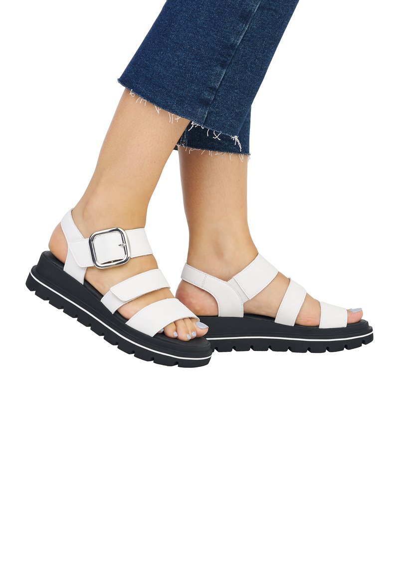 White strappy sandals with a buckle detail, black outsole, and a thick sole. Worn with cropped denim jeans featuring frayed hems.