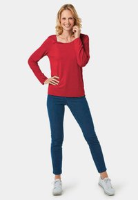 Rotes Langarmoberteil mit quadratischem Ausschnitt, kombiniert mit blauen Slim-Fit-Jeans und weißen Turnschuhen. Glattes, weiches Material mit lässigem Schnitt.