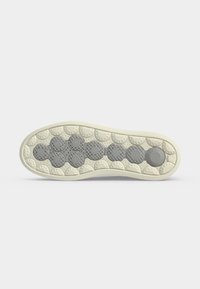 Semelle d'une chaussure avec des grips texturés gris circulaires disposés en un motif sur fond beige contre une surface blanche unie.