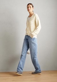 Cardigan di colore crema con un design a righe testurizzato, abbinato a jeans di denim azzurro chiaro e scarpe grigie, in piedi su un pavimento in legno.