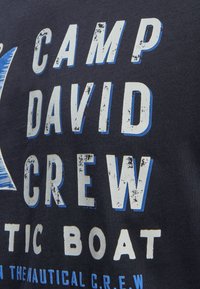 Navy blue t-shirt featuring white and blue distressed text reading "CAMP DAVID CREW" and "NAUTIC BOAT." Printed graphic with a triangular design.