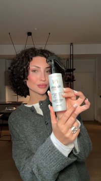 Une femme aux cheveux bouclés tient un vaporisateur étiqueté "KEATIN" dans une cuisine moderne, présentant le produit de près à la caméra.