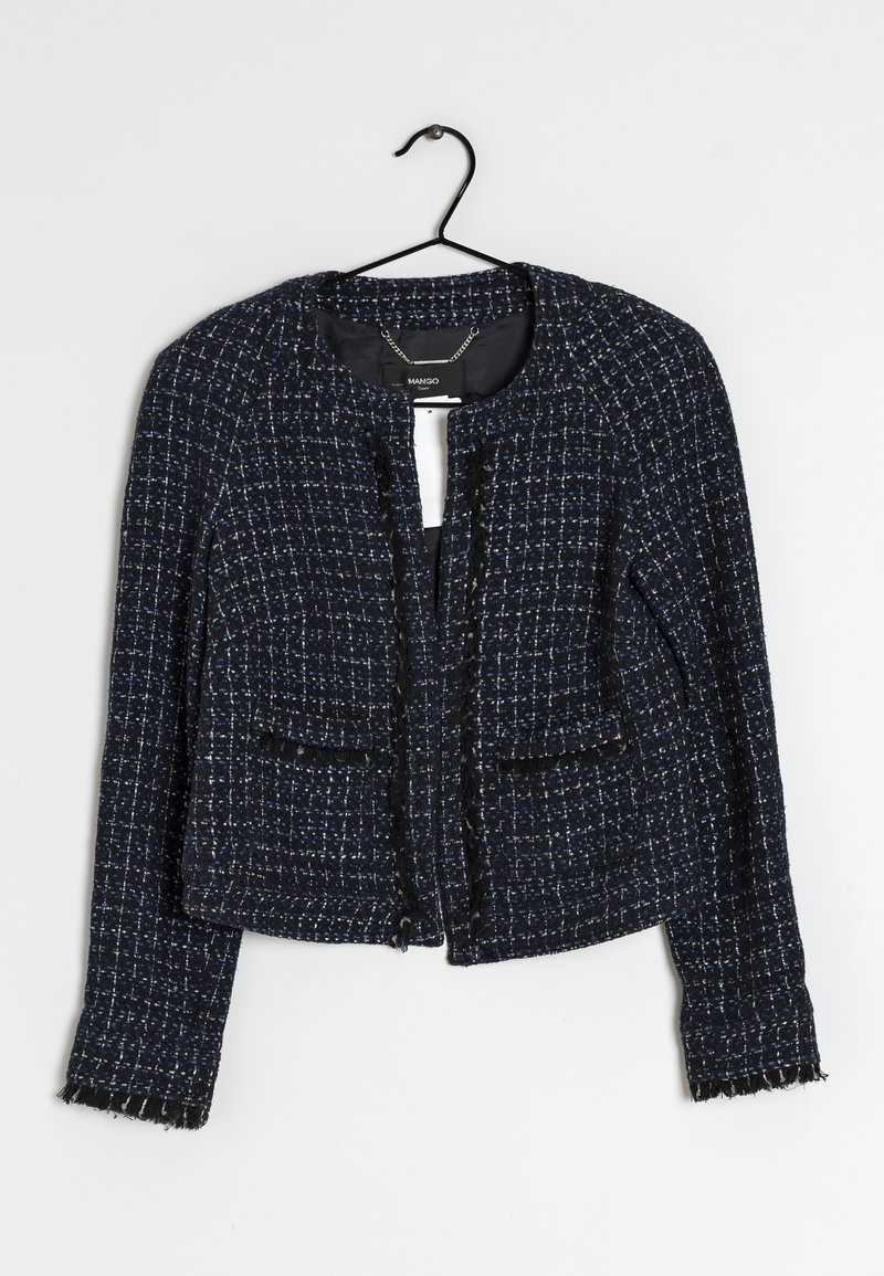 Veste courte en tweed bleu foncé et noir avec manches longues et bords frangés suspendue à un cintre noir sur fond blanc.