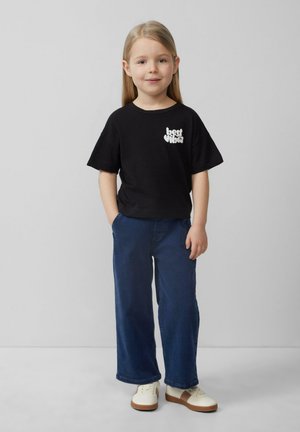 Czarny bawełniany t-shirt z grafiką "najlepsze wibracje" na piersi, w zestawie z granatowymi szerokimi spodniami i białymi trampkami z brązowymi akcentami.