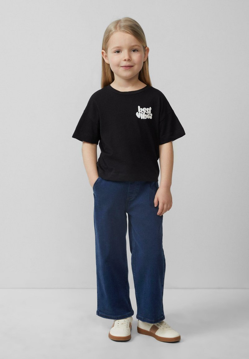 Czarny bawełniany t-shirt z grafiką "najlepsze wibracje" na piersi, w zestawie z granatowymi szerokimi spodniami i białymi trampkami z brązowymi akcentami.
