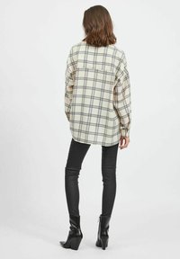 Woman with shoulder-length hair wearing oversized cream and gray plaid shirt, black fitted pants, and black heeled ankle boots, standing facing away.