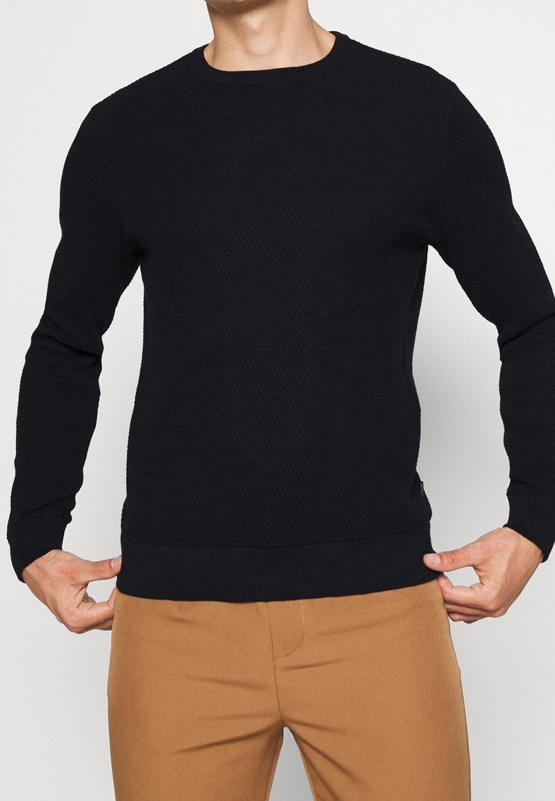 Homme portant un pull ajusté noir texturé à manches longues et un pantalon marron clair, tenant les bords inférieurs du pull.