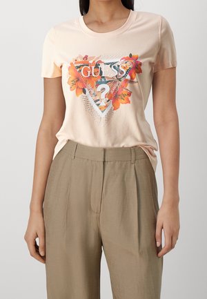 Kvinde iført en lyserød Guess t-shirt med blomstertrianglelogo og beige højhalsede plisserede bukser mod en ensfarvet baggrund.