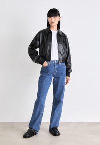 Kvinne som står med hånden i lommen, iført svart skinnjakke, hvit t-skjorte med logo, blå jeans og svarte slip-on sko mot en ensfarget vegg.