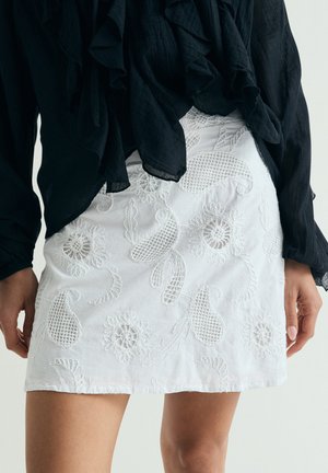Minifalda blanca bordada con motivos florales y de paisley, combinada con un top negro con volantes y mangas largas, manos relajadas a los lados.