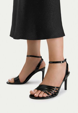 Stopy w czarnych lakierowanych sandałach na wysokim obcasie z paskami wokół kostek, zestawione z czarną teksturowaną spódnicą.