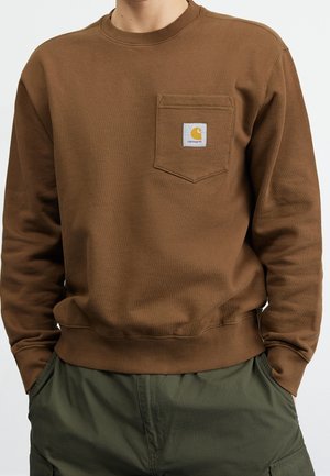 Brązowa bluza z długim rękawem z kieszenią na piersi z logo Carhartt, noszona z zielonymi spodniami typu cargo, ręce w kieszeniach.