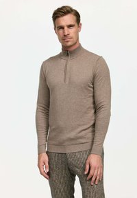 Brūna, adīta ceturtdaļas zips džemperis ar stāvošu apkakli un garām piedurknēm. Mīksta tekstūra, pieguļoša forma un ribota apakšmala.