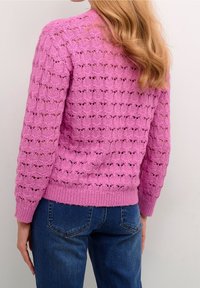 Rosa gestrickter Pullover mit offener Spitzenoptik, gerippten Bündchen und Saum. Der Rücken zeigt eine entspannte Passform, kombiniert mit dunkelblauen Jeans.