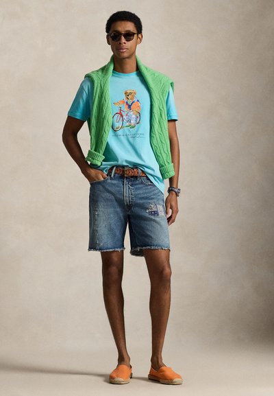 Άνδρας με γυαλιά ηλίου, μπλε πουκάμισο με σχέδιο, πράσινο πουλόβερ περασμένο πάνω από τους ώμους, τζιν σορτς και πορτοκαλί παπούτσια χωρίς κορδόνια, στέκεται μπροστά σε ουδέτερο φόντο.