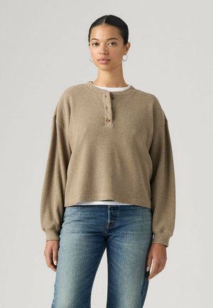 Giovane donna con capelli scuri, indossa una maglia beige a maniche lunghe con bottoni e jeans blu, in piedi davanti a uno sfondo chiaro e semplice.
