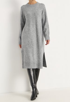 Mujer con un vestido midi de punto gris de manga larga con abertura lateral y botas de cuero negras hasta la rodilla, sobre un fondo blanco.