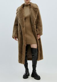 Langer brauner Kunstfellmantel mit breitem Kragen, kombiniert mit einem gerippten beigen Kleid und schwarzen, knielangen Stiefeln mit dicken Absätzen.