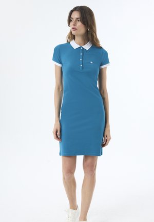 Kvinde iført en blå polo kjole med hvid krave og ærmekanter, står og kigger lidt til siden mod en hvid baggrund.