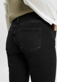 Jeans de mezclilla negra con un diseño ajustado, costuras sutiles y bolsillos parche estándar. La textura es suave con un ligero estiramiento.