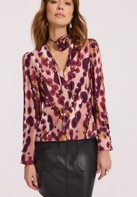 Kobieta ubrana w bluzkę w kwiaty z długim rękawem w odcieniach różu i fioletu, dopasowany kwiatowy choker oraz czarną skórzaną spódnicę.