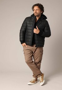 Doudoune noire avec fermeture éclair, poignets côtelés et capuche. Pantalons cargo marron avec poches latérales. Baskets montantes marron et blanches.