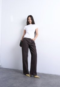 Γυναίκα που στέκεται μπροστά σε λευκό τοίχο, φοράει λευκή μπλούζα, σκούρο παντελόνι φαρδιάς γραμμής, μπεζ τακούνια και κρατάει μια μικρή μαύρη τσάντα στο πλάι της.