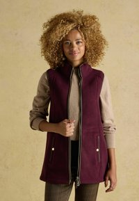 Burgunderfarbene ärmellose Weste mit hohem Kragen, Reißverschluss auf der Vorderseite und zwei Seitentaschen; darüber eine beige Strickjacke mit gerippten Ärmeln.