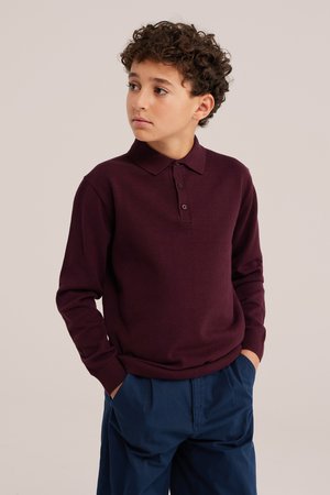 Garçon aux cheveux bouclés portant un polo bordeaux à manches longues et un pantalon bleu, regardant vers sa gauche avec les mains dans les poches.