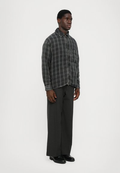 Camicia a quadri grigia con bottoni e taschino frontale, abbinata a pantaloni larghi scuri e scarpe nere. Design semplice, vestibilità rilassata.