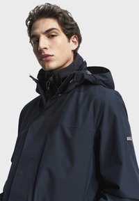 Marinblå vattentät jacka med dragkedja och tryckknappar, som har en hög krage, justerbar huva och logotypapplikation på ärmen.