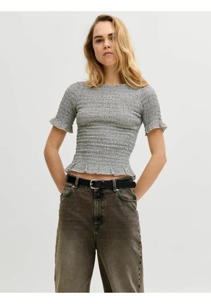 Blonde Frau trägt ein eng anliegendes graues, strukturiertes Kurzarmshirt und hoch taillierte dunkelbraune Jeans mit schwarzem Gürtel, steht mit Händen in den Taschen.