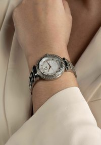 Orologio argentato con lunetta intrecciata, quadrante bianco a sole, numeri romani e bracciale in metallo, presentato su uno sfondo di tessuto chiaro.