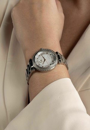 Orologio argentato con lunetta intrecciata, quadrante bianco a sole, numeri romani e bracciale in metallo, presentato su uno sfondo di tessuto chiaro.