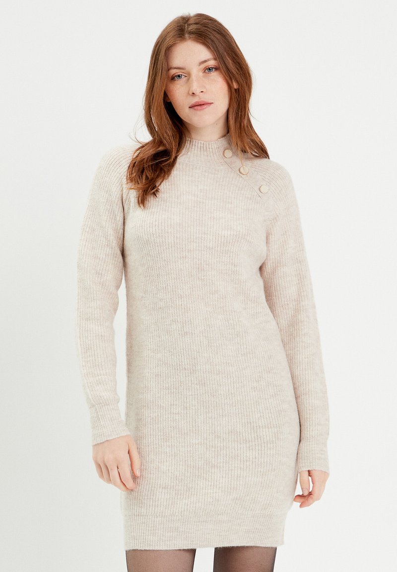 Vestido de punto acanalado beige con mangas largas, cuello alto y tres botones decorativos en el hombro izquierdo. Textura suave, silueta ajustada.