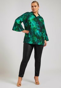 Blusa de manga larga, verde y negra con un patrón, con cuello y puños acampanados, combinada con pantalones de cuero negros ajustados y sandalias doradas de tacón.